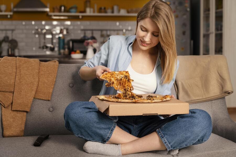 Mulher sentada no sofá da sua casa comendo uma pizza.